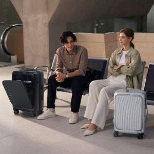 Modern travelers in stylish attire seated at airport terminal, OIWAS SMOOTH matte-finish carry-ons at their feet.